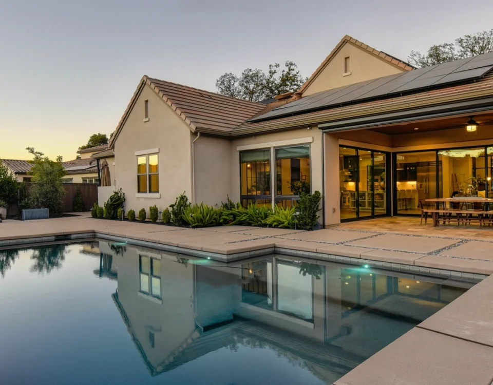 Backyard pool at dusk beside a house with solar panels, open glass doors, and a lit patio dining area
